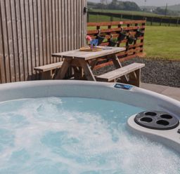 An outdoor hot tub with bubbling water next to a wooden picnic table and a fence with planters at Parys Pod in Amlwch