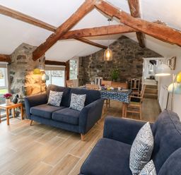 A living room with blue sofas wooden beams stone walls and a dining table with chairs at Y Bwthyn in Dyffryn Ardudwy