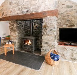 A living room with a stone fireplace with a wood stove a blue chair a small wooden table with teacups a flat screen TV and a basket with a blanket at Y Bwthyn in Dyffryn Ardudwy