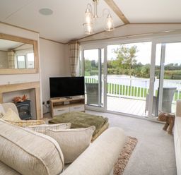 A living room with beige sofas a television a fireplace and glass sliding doors leading to a deck overlooking a pond at 10 Cheviot View in Haggerston near Lowick