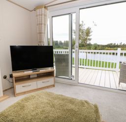 A living room with a television on a wooden stand and glass sliding doors opening to a balcony overlooking water at 10 Cheviot View Haggerston near Lowick