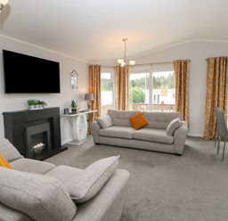 A living room with two gray sofas orange cushions a black fireplace a wall-mounted TV and a dining table by windows with orange curtains at Mildred's Lodge in Felton