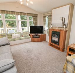 A living room with a grey sofa a fireplace a TV on a wooden corner unit and window seating with green plaid cushions at Hedgehog Retreat in Felton