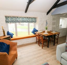 A dining area with a wooden table set for four next to a window seat and wicker chair at Bluebell Cottage in Jameston near Manorbier