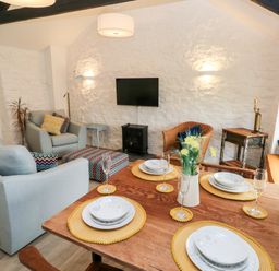 A dining and living area with a wooden table set with plates and glasses and sofas with cushions at Bluebell Cottage in Jameston near Manorbier