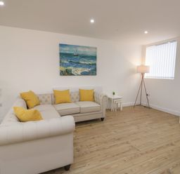 A living room with a beige sofa with yellow pillows a sea painting on the wall a floor lamp a wall-mounted television and wooden flooring at The Church in Holyhead