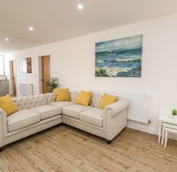 A living room with a beige sectional sofa with yellow cushions a painting of sailboats on the sea and nested white side tables at The Church in Holyhead