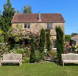 A garden with benches flowers and bushes in front of a stone house with tiled roof at Ham cottage East Lambrook near South Petherton