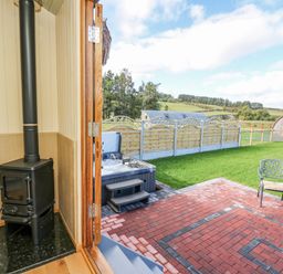 A small wood-burning stove inside a room with an open door leading to a patio with a hot tub a chair and a grassy yard at Poppy in St Harmon near Rhayader