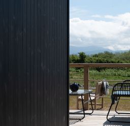 A wooden deck with chairs a table and a teapot overlooking green bushes and mountains at Galwad y Mor in Niwbwrch newborough