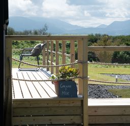 A wooden deck with a chair and a potted plant with a sign near greenery and distant mountains at Galwad y Mor in Niwbwrch newborough
