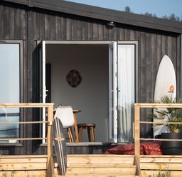 A wooden deck in front of a dark house with open double doors showing a table and chairs inside a surfboard and skateboard on the deck with a potted plant at Galwad y Mor in Niwbwrch newborough