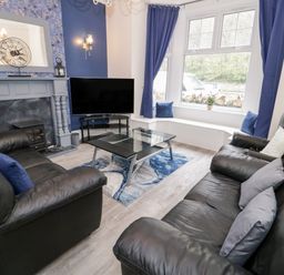 A living room with black leather sofas a glass coffee table a fireplace and a large window with blue curtains at Gwyder in Penmaenmawr