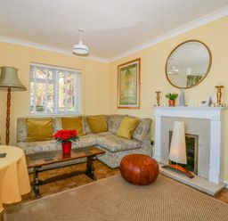 A living room with a patterned sofa and yellow cushions a wooden coffee table with a red flower pot a fireplace with a large round mirror above it and a woven rug on the floor at See The Sea in Hunstanton