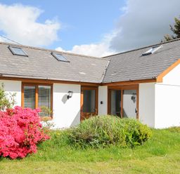 A white house with wooden framed windows and doors pink flowers and green bushes in front at Lambley View in Bude