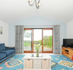 A living room with a blue couch a white coffee table a television on a wooden stand and sliding glass doors opening to a garden at Lambley View in Bude