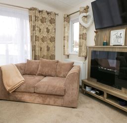 A living room with a brown sofa beige curtains a mounted television and a wooden cabinet with decorative items at 3 Yealands Carnforth