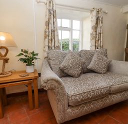 A living room with a patterned sofa with cushions next to a wooden table with a lamp and a potted plant at 5 Packhorse in Purton