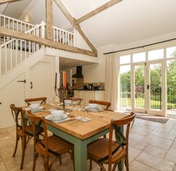 A dining area with a wooden table set for six next to large glass doors and a staircase with an open loft at The Hayloft in Stockland Honiton