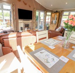 A living room and dining area with leather sofas a dining table with placemats and a vase of roses a wall-mounted television and windows at Mallard Lodge in Carnforth