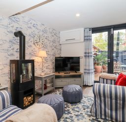 A living room with striped armchairs two ottomans a wood stove and large glass doors at Tower End Cottage in Melton