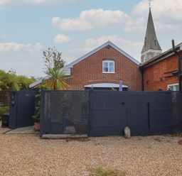 A courtyard with a gravel ground and a dark gate in front of a brick building with a church steeple in the background at Tower End Cottage in Melton