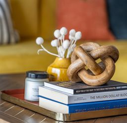 A wooden coffee table with a tray holding books a candle a yellow vase with white decorative flowers and a wooden knot sculpture with a yellow sofa and cushions in the background at Porth Beach Villa in Porth near Newquay