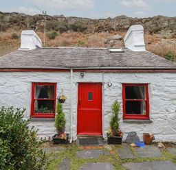 A stone cottage with red door and window frames with plants and rocky hill behind at Quarry Cottage in Blaenau Ffestiniog