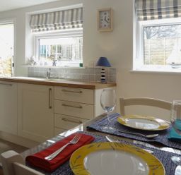 A kitchen with a dining table set for two near two windows with striped blinds and a green door at Fiddlesticks Cottage in Beaminster