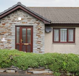 The exterior of a single-story house with a stone entrance door and a window with curtains at St Albans in Boscastle