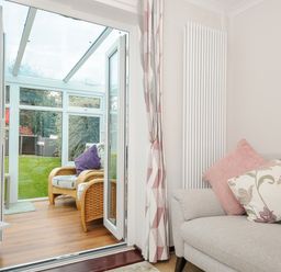 A living room with a gray sofa and floral cushions next to glass doors leading to a conservatory with wicker chairs at St Albans in Boscastle