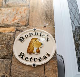 A ceramic sign with a dog's head and the words Bonnies Retreat mounted on a stone wall next to a white door with diamond-patterned glass at Bonnies Retreat in Amble