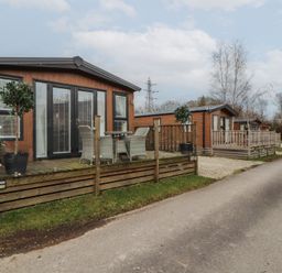 A row of wooden cabins with small fenced porches and outdoor seating along a paved road at 24 Sherwood in Carnforth