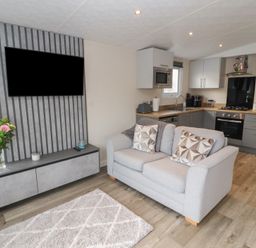 A living room with a grey sofa, a wall-mounted TV, flowers on a cabinet, and a kitchen with appliances at 24 Sherwood in Carnforth