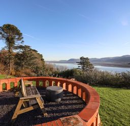 An outdoor seating area with a wooden bench and round table overlooking a river with hills and trees at Bryn Mel Manor Straits View in Menai Bridge
