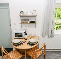 A small dining area with a round wooden table set for two with dishes and wine next to a microwave under shelves with dishes near a window with grey curtains at Ty Rhydd in Newport Pembrokeshire