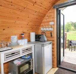 A small kitchen area with a refrigerator microwave kettle plates bowls glasses and mugs with an open door showing a barbecue outside The Pod at Bank House Farm The Blythe near Hixon