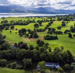 An aerial view of green fields with scattered trees and some houses near a body of water with mountains in the background at Ty Bach - Beaumaris in Beaumaris