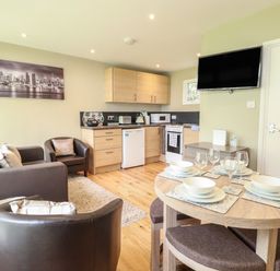 A small living area with a sofa, armchairs, a kitchen with wooden cabinets, a dining table set with plates and glasses, and a wall-mounted TV at 107 Kings Chalet Park in Cromer