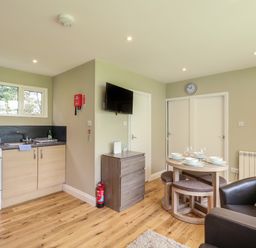 A kitchen area with stove and sink next to a dining table set with plates and glasses a wall-mounted TV and a brown armchair at 107 Kings Chalet Park in Cromer