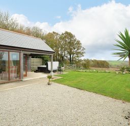 An outdoor area with a gravel patio, grassy lawn, small palm tree, wooden building with large windows, hot tub and metal garden table at Pol Glas Cabin in Churchtown near Mullion