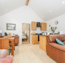 A living area with a brown leather sofa, wooden dining table with chairs, and a kitchenette at Pol Glas Cabin in Churchtown near Mullion