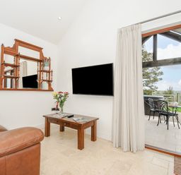 A living room with a brown leather sofa a wooden coffee table a wall-mounted tv and a large mirror next to glass doors leading to an outdoor seating area with a view at Pol Glas Cabin in Churchtown near Mullion