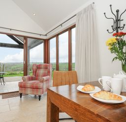A dining table with croissants cups and teapot next to armchair and large windows at Pol Glas Cabin Churchtown near Mullion