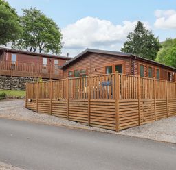 A wooden lodge with a fenced deck and gravel driveway surrounded by trees at Hollytree Lodge Beckside 1 Limefitt Park near Windermere