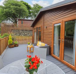 An outdoor deck with wooden railing featuring a round metal table with a red flower pot and chairs beside a wooden cabin with glass doors at Hollytree Lodge in Beckside 1 Limefitt Park near Windermere