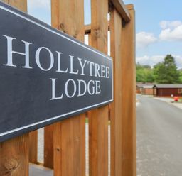 A wooden fence with a Hollytree Lodge sign and a road with wooden buildings in the background at Hollytree Lodge Beckside 1 Limefitt Park near Windermere