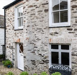 Exterior view of a stone cottage with white framed windows a white door and a metal bench at Honey Cottage in Port Isaac