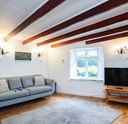 A living room with a grey sofa a wooden TV stand with a television a window and wooden ceiling beams at Honey Cottage in Port Isaac