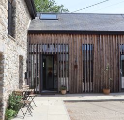 An outdoor patio area with a wooden table and chairs next to a stone building and a wooden slatted facade at 3 Caddaford Barns in Buckfastleigh